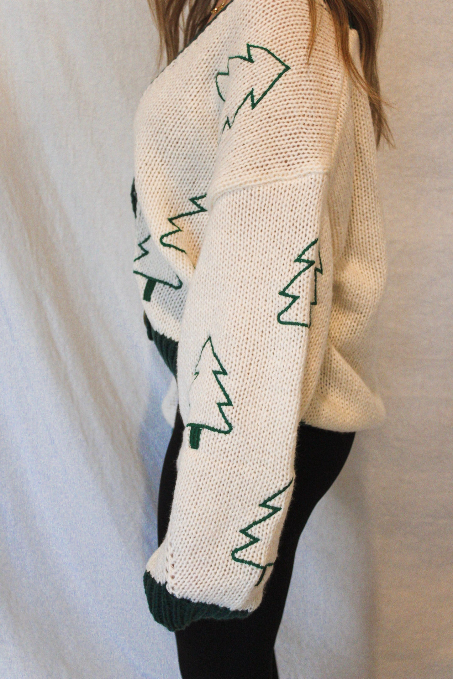 Cream-colored sweater with green tree patterns worn by a person against a plain background