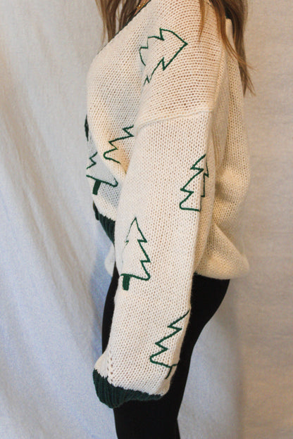 Cream-colored sweater with green tree patterns worn by a person against a plain background