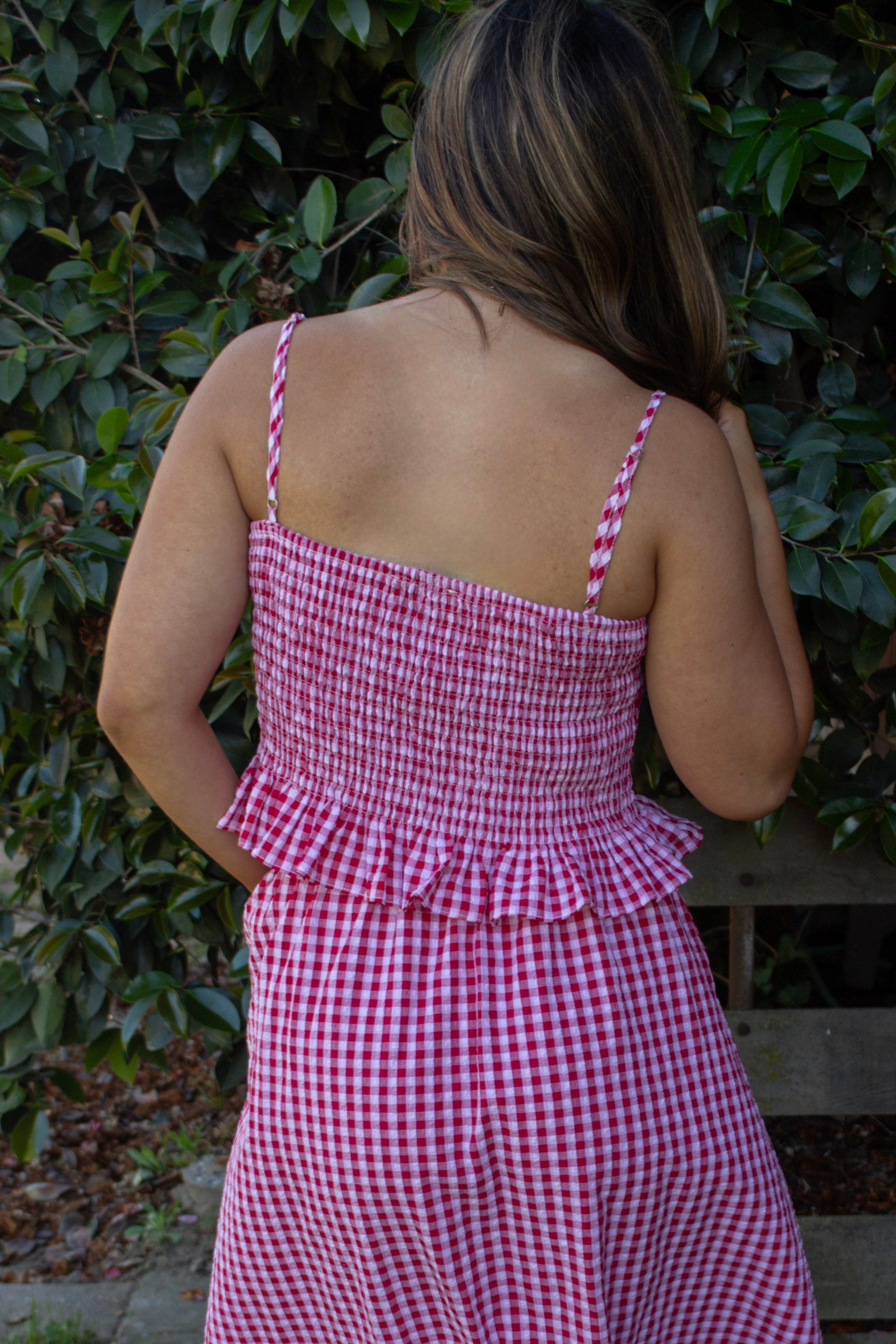 Person wearing a pink checkered dress standing outdoors with greenery in the background