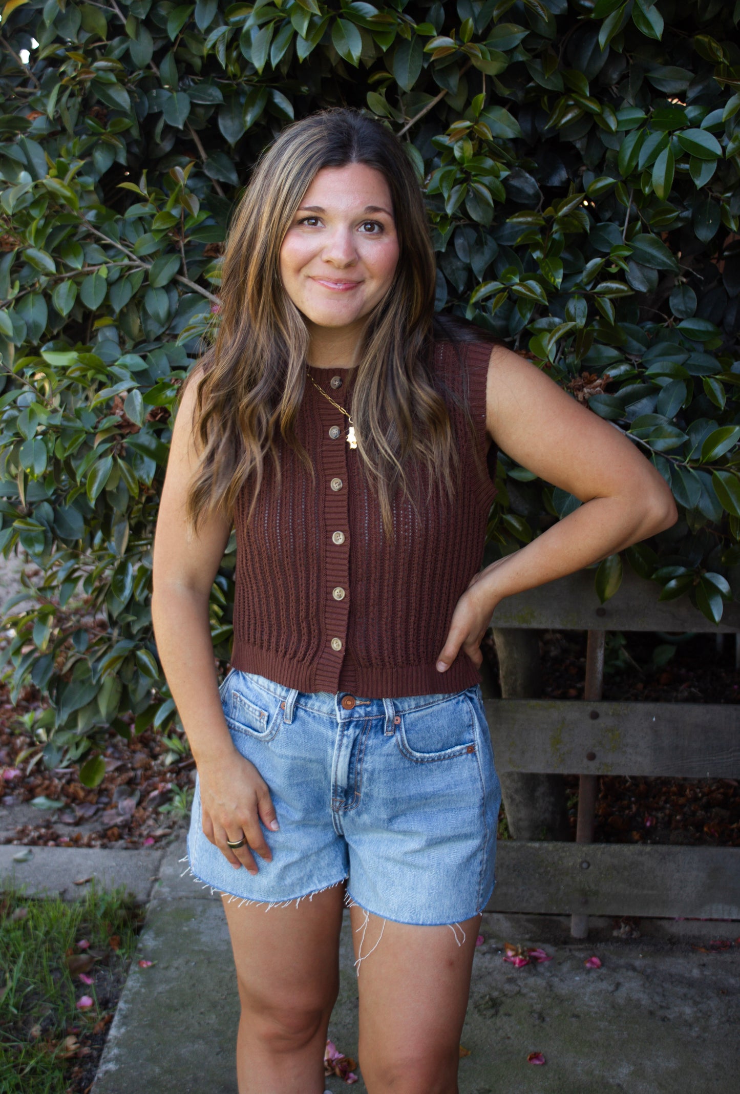 Woman wearing a brown sleeveless sweater and denim shorts standing in front of green foliage.