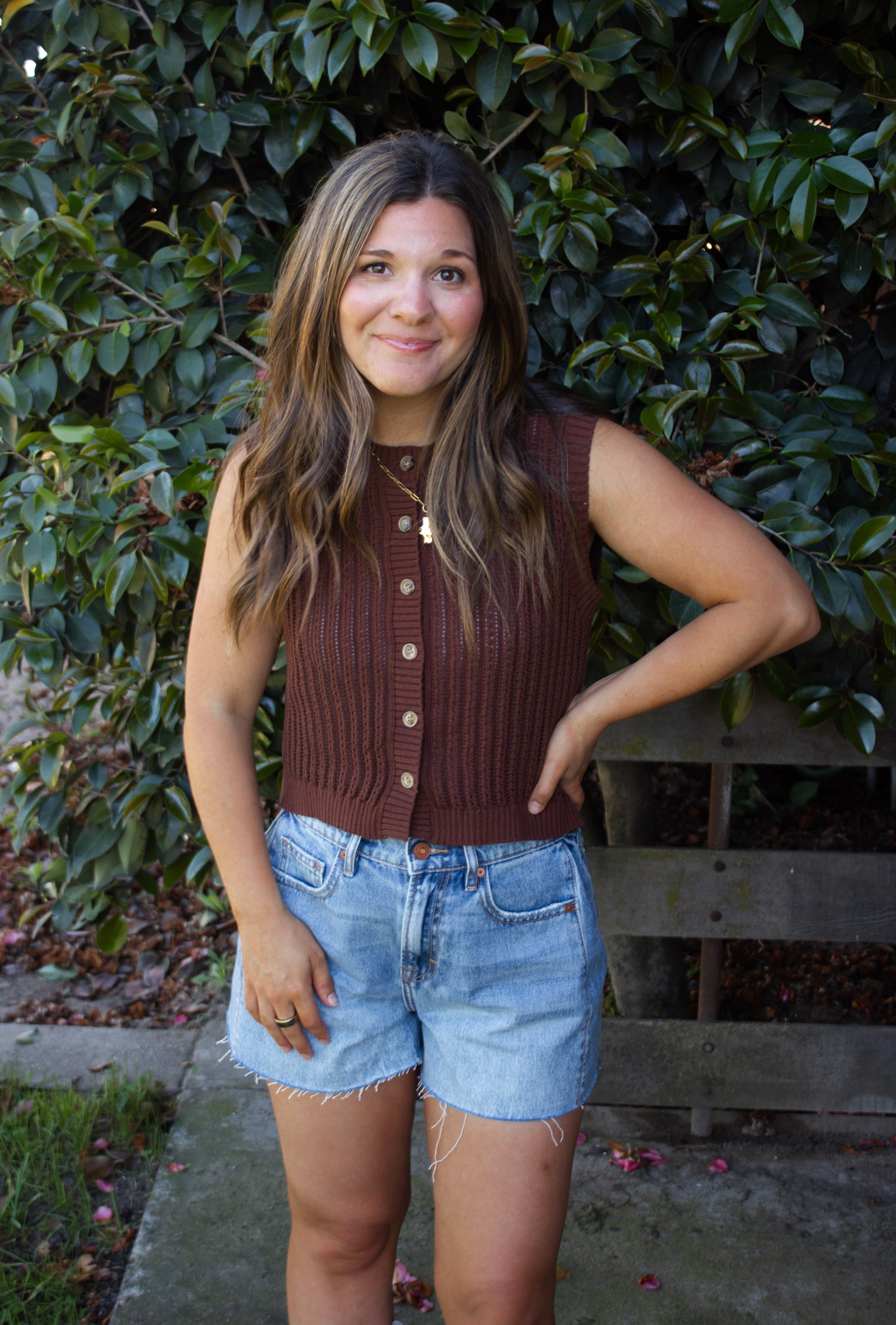 Woman wearing a brown sleeveless sweater and denim shorts standing in front of green foliage.