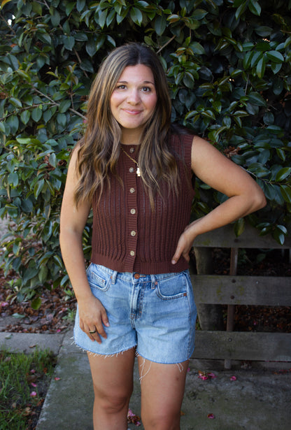 Woman wearing a brown sleeveless sweater and denim shorts standing in front of green foliage.