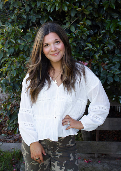 Woman wearing a white blouse with lace details and camouflage pants standing in front of green foliage.
