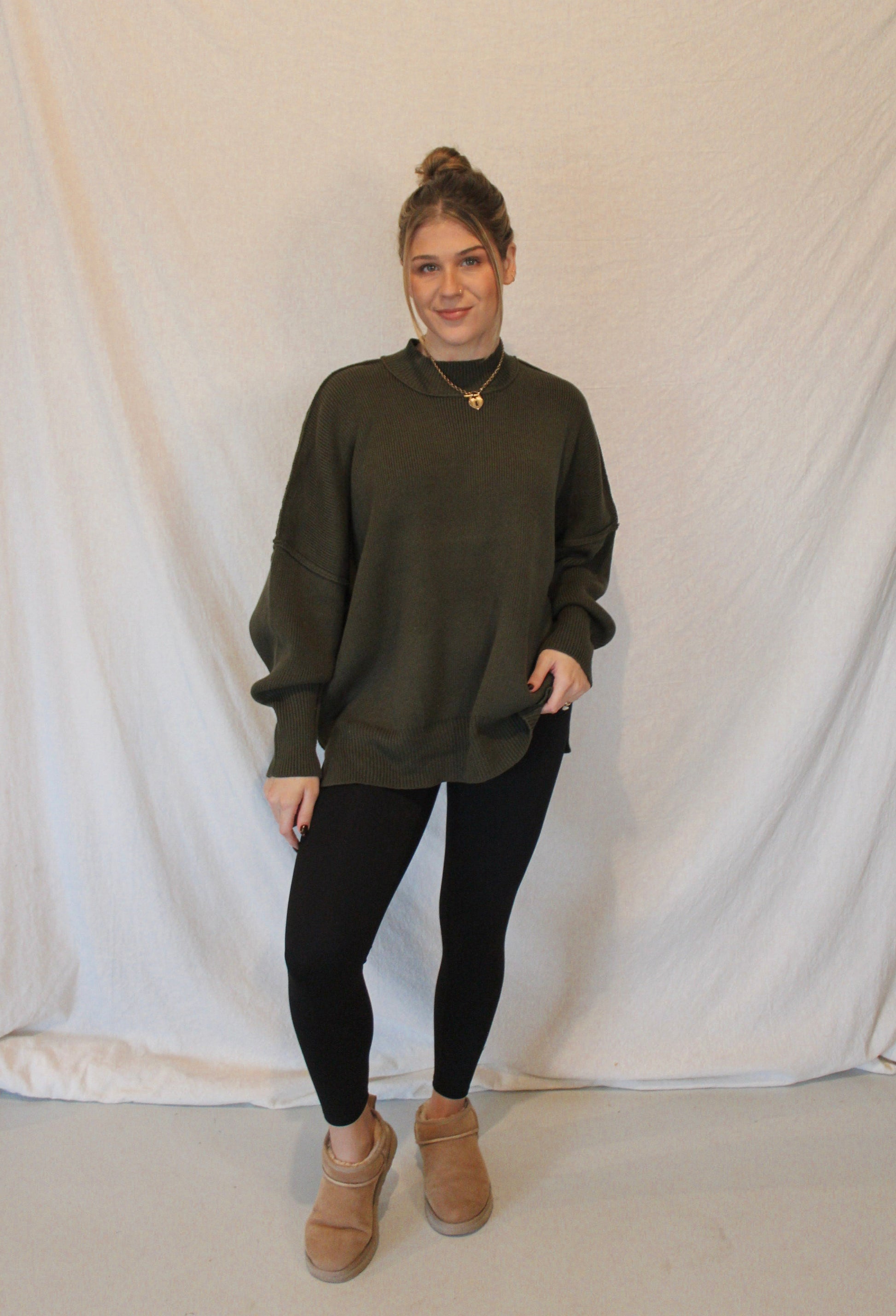 Person wearing an olive green sweater, black leggings, and brown boots against a white backdrop