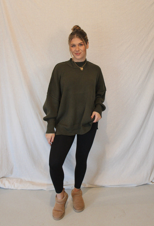 Person wearing an olive green sweater, black leggings, and brown boots against a white backdrop
