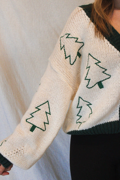 White sweater with green tree patterns worn by a person on a light gray background