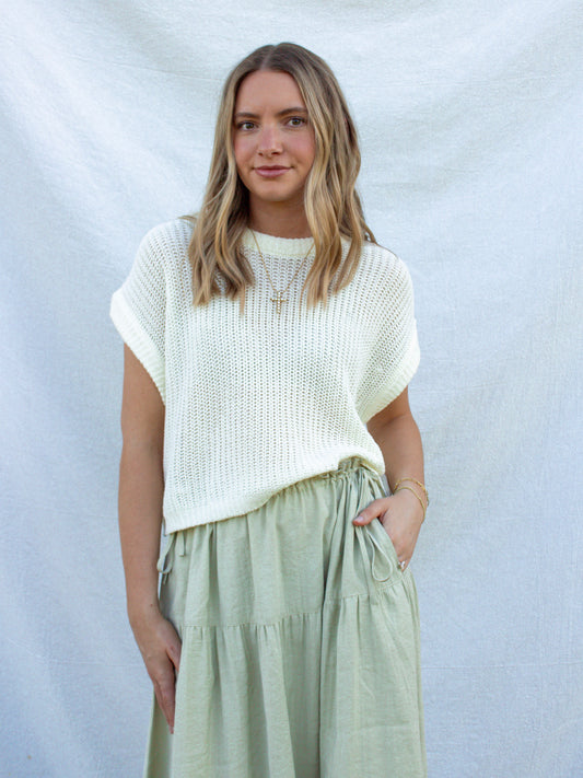 Woman wearing a white knit top and light green pants against a plain background