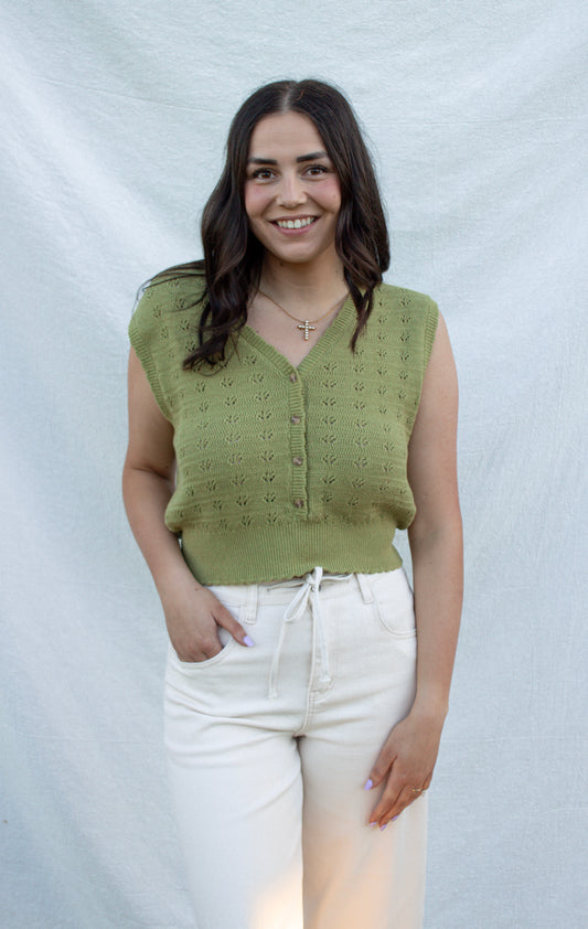 Woman wearing a green knitted vest and white pants against a plain background