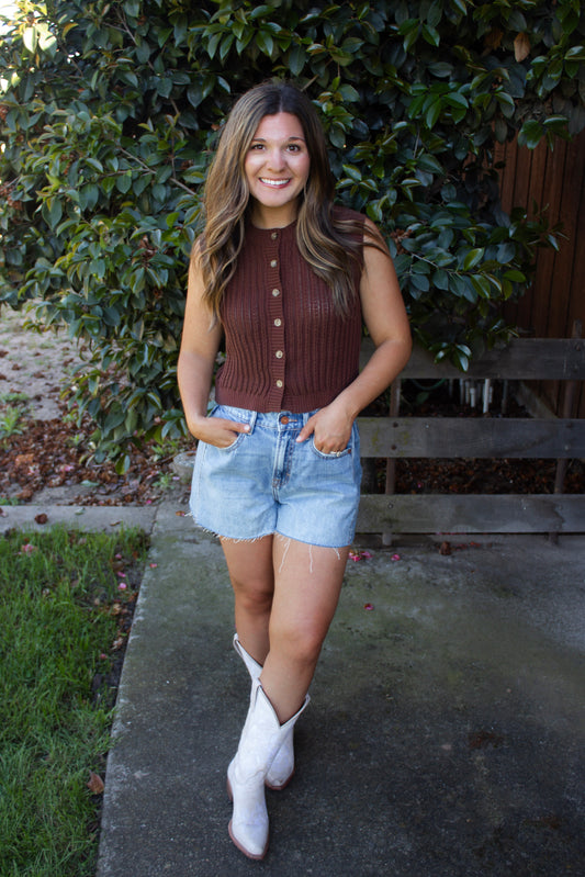 Woman in a brown ribbed sweater, denim shorts, and white cowboy boots standing outdoors.