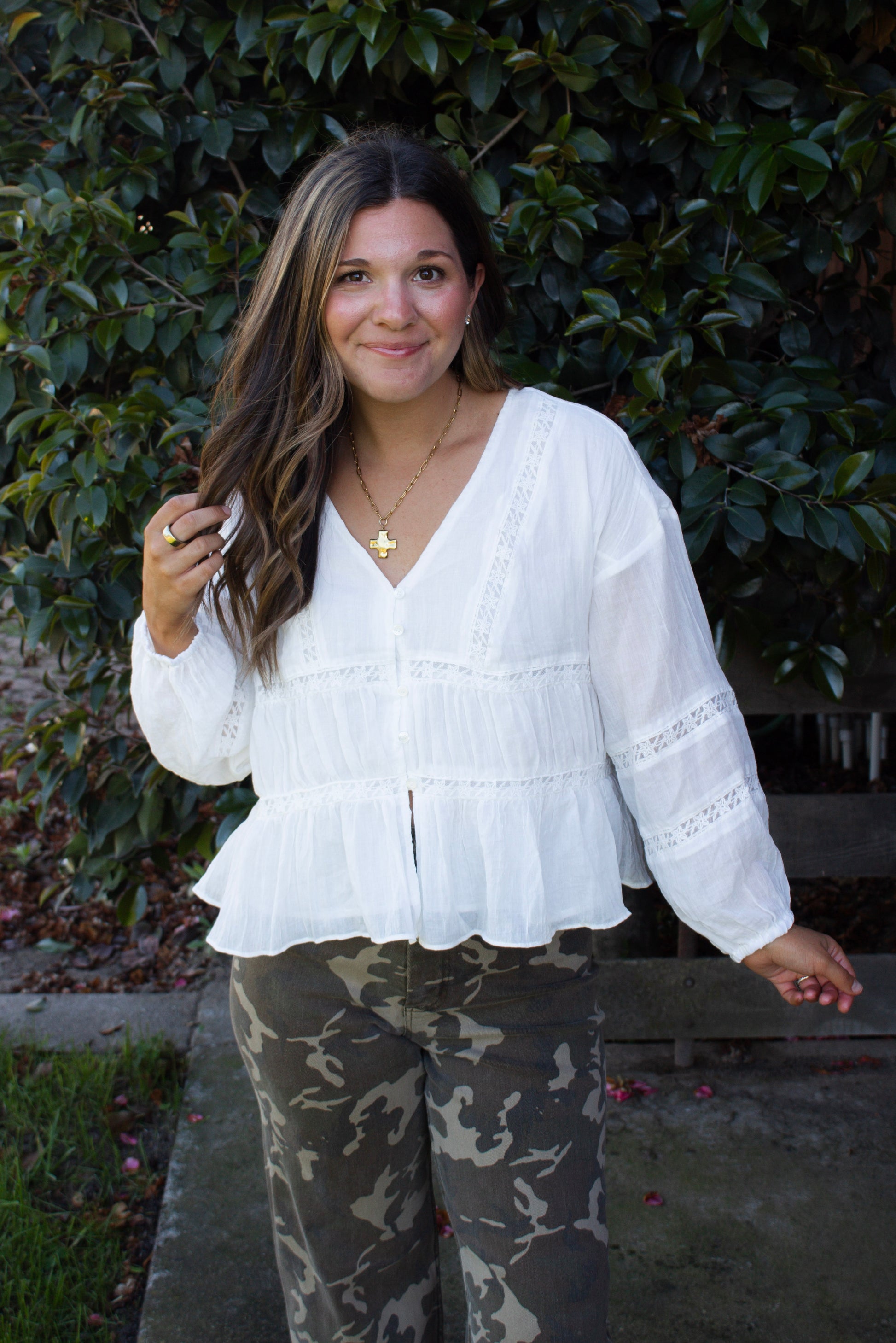 Woman wearing a white blouse and camouflage pants standing against a green bush.