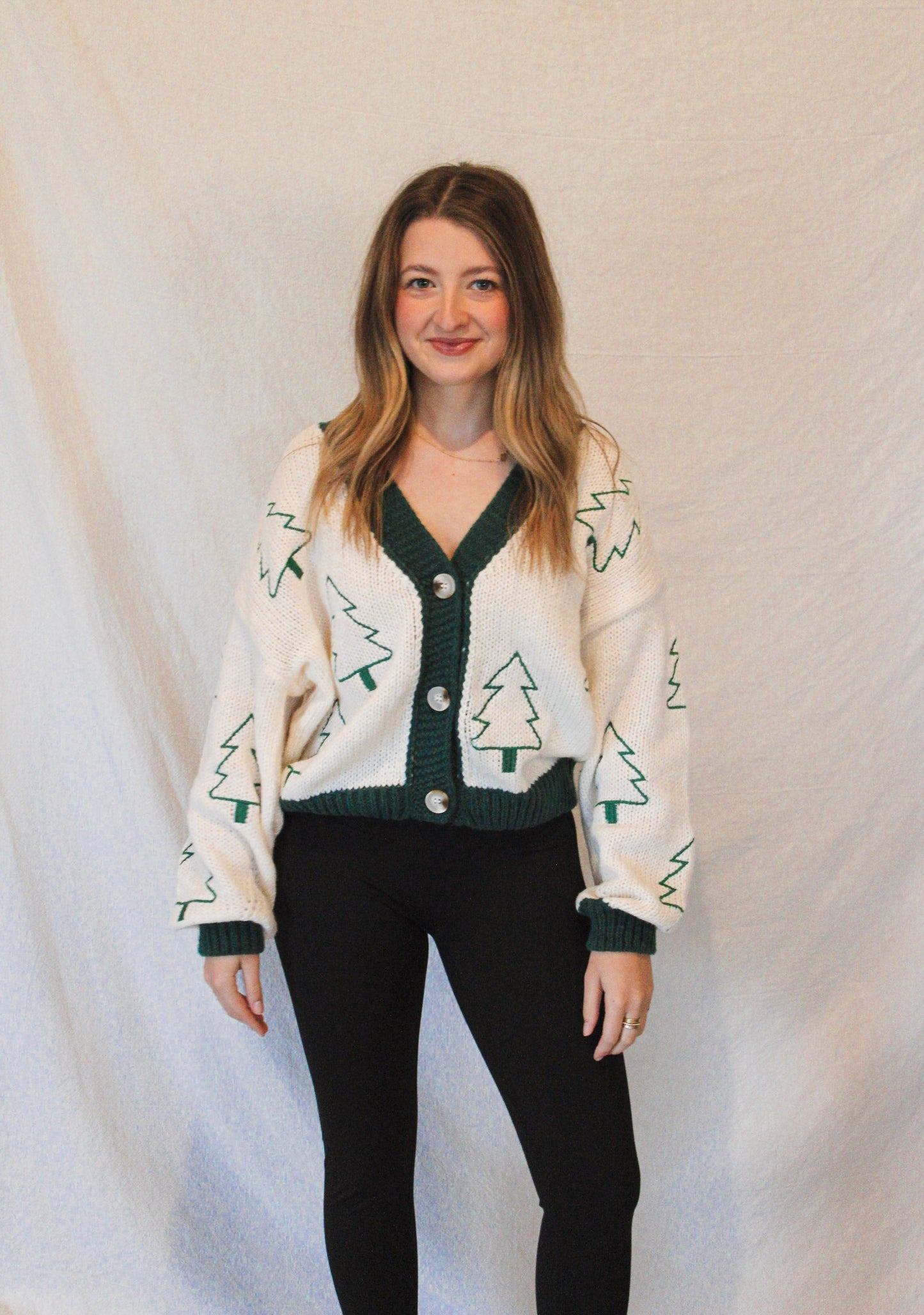 Woman wearing a cream cardigan with green pattern and black pants against a plain background