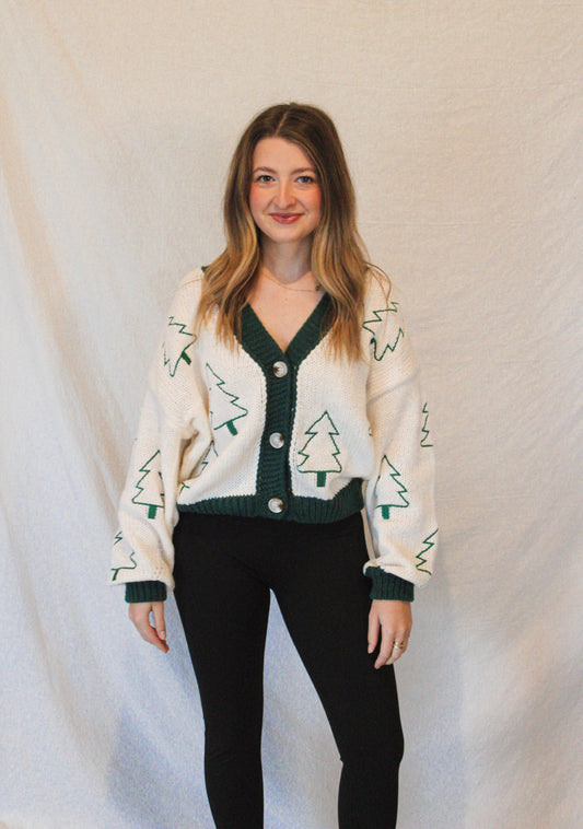 Woman wearing a cream cardigan with green pattern and black pants against a plain background