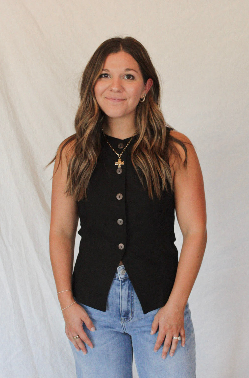 Woman wearing a black sleeveless top with buttons and light blue jeans against a plain background