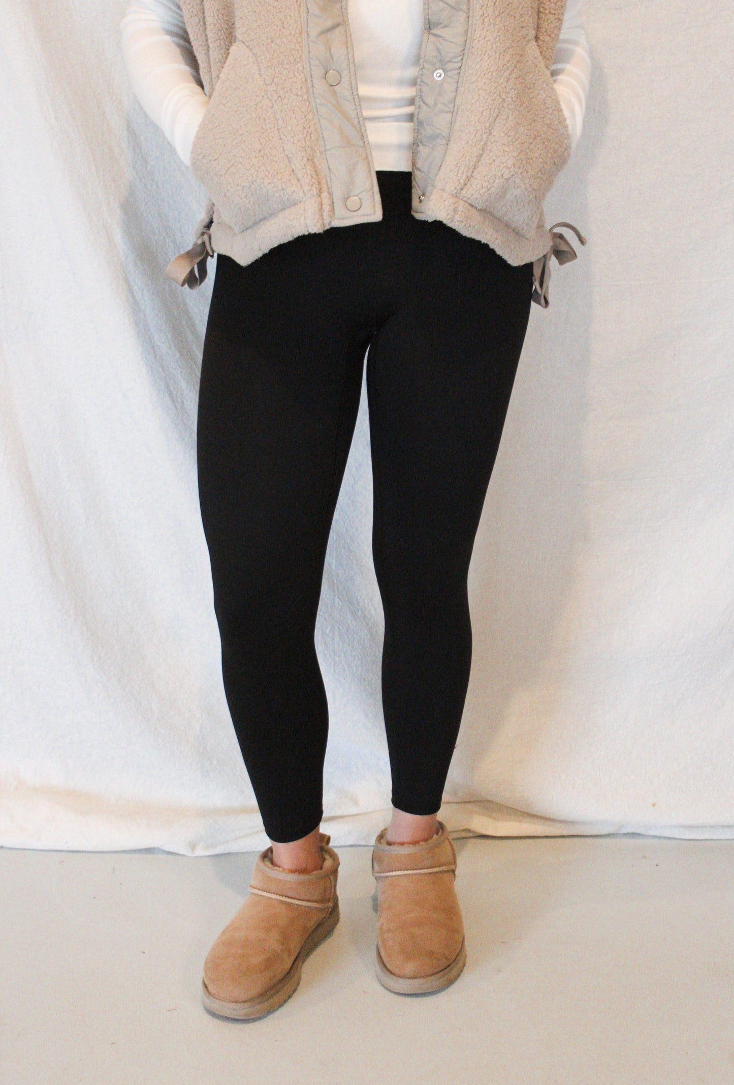 Person wearing black leggings, beige jacket, white shirt, and tan shoes on a plain background