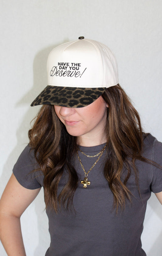 Person wearing a white cap with leopard print visor and 'Have the Day You Deserve!' text.